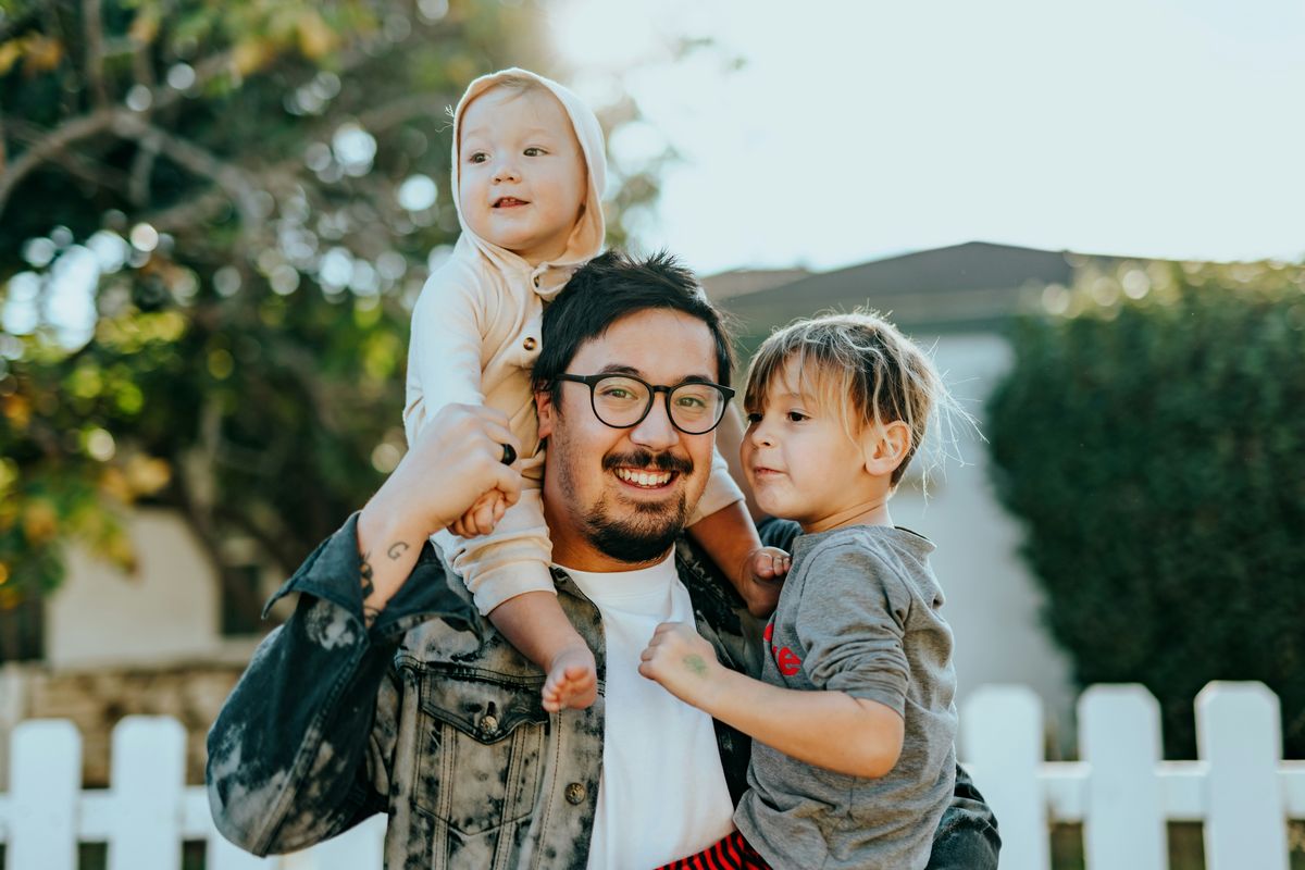 A smiling dad outdoors with his two young sons, one on his shoulders and one in his arms